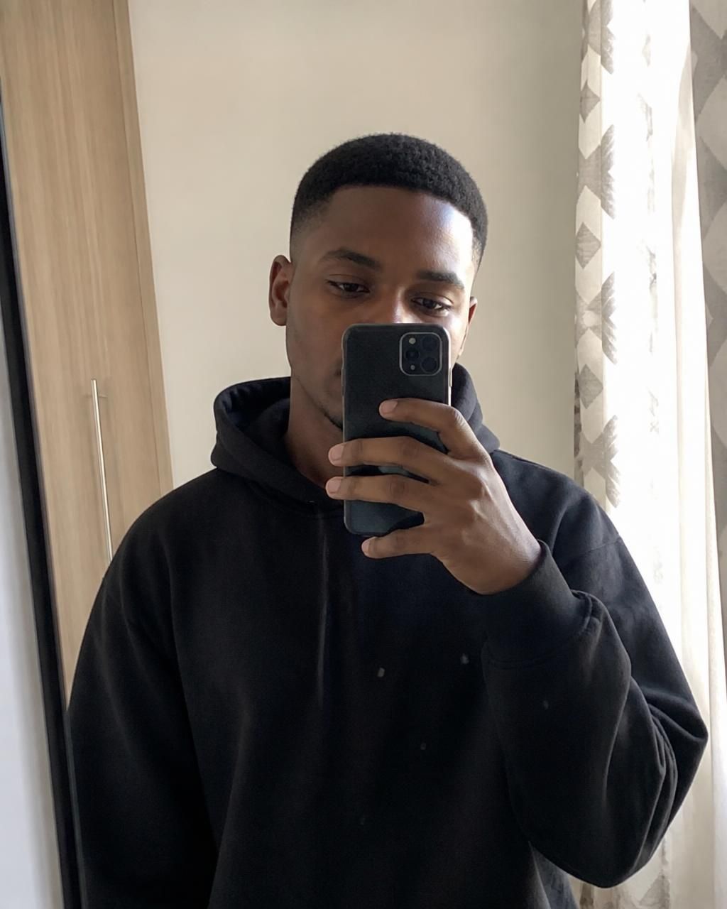 Mirror selfie of a man wearing a plain black hoodie in soft indoor light