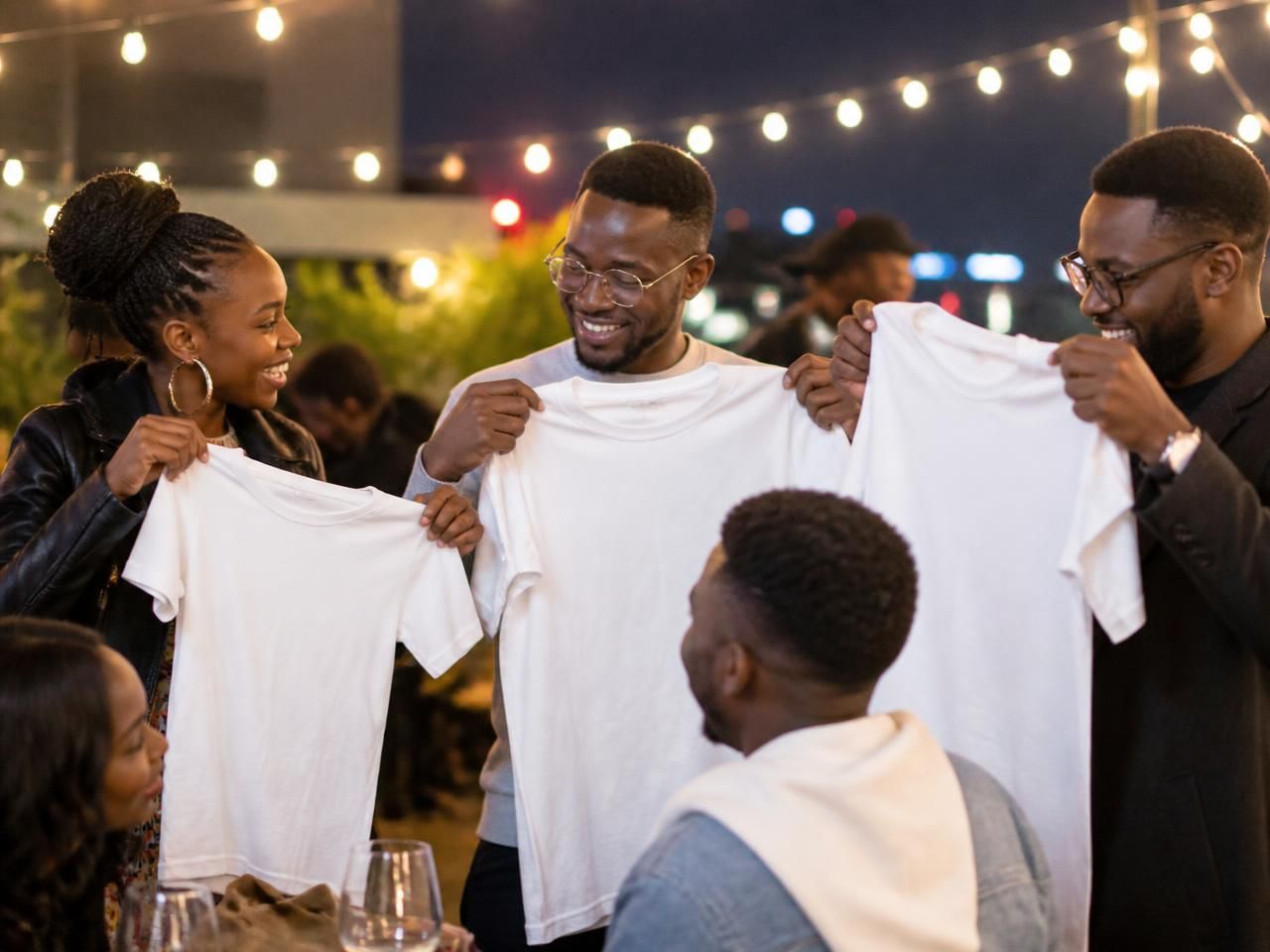 Friends holding up matching white T-shirts under string lights at an evening celebration